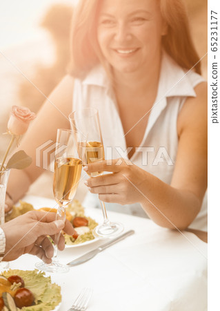 Happy woman smiles holding glass of white wine. Mature couple have event outdoors at beach cafe. Selective focus on human hands with glasses of wine or champagne in foreground. Toned image 65231177