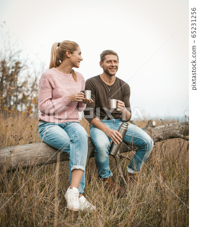 Smiling couple of tourists drink tea and talk while sitting on fallen tree among an autumn nature outdoors Smiling couple of tourists drink tea and talk while sitting on fallen tree among an autumn nature outdoors 65231256