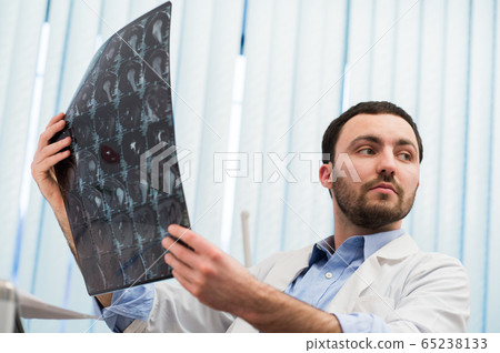 Closeup portrait of intellectual man healthcare personnel with white labcoat, looking at brain x-ray radiographic image, ct scan, mri, clinic office background. Radiology department 65238133