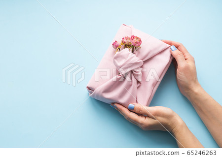 Female hands holding a holiday gift packed fabric in the manner of Furoshiki on a fashionable blue background. Female hands holding a holiday gift packed fabric in the manner of Furoshiki on a fashionable blue background. 65246263