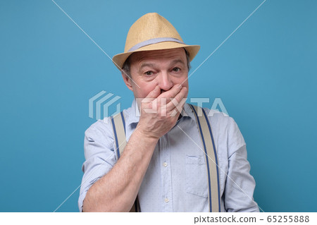 Stunned elderly man in summer hat covers mouth with hands, being surprised 65255888