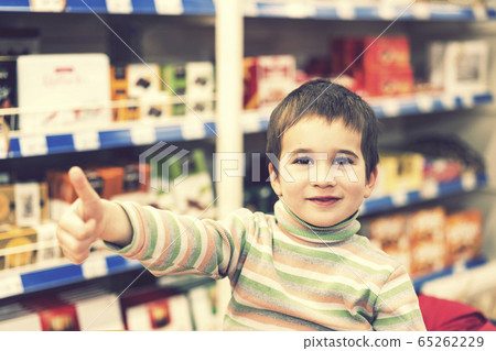 Happy boy 4 years in a supermarket on the background of shelves with chocolate. The boy showed that everything is ok. toned 65262229