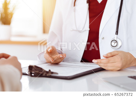 Unknown woman-doctor and patient discussing something while sitting at the table in sunny clinic, close-up Unknown woman-doctor and patient discussing something while sitting at the table in sunny clinic, close-up 65267372