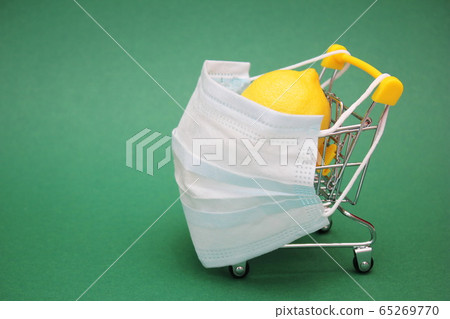 A medical mask is placed on a shopping cart containing a fresh lemon. Green background. 65269770