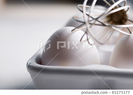Ceramic box with natural organic farm eggs and metal whisk on a wooden kitchen table. 65273177