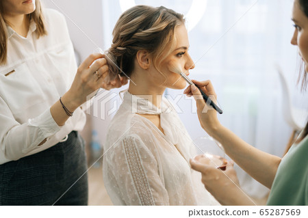 Side view of make-up artist putting rouge on woman's cheek using brush 65287569