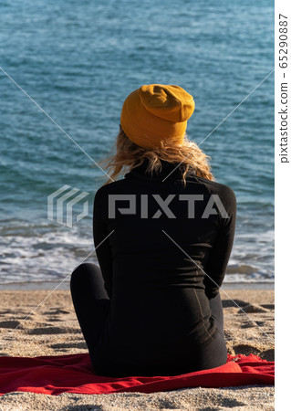 A girl with blond curly hair in black sport jacket and yellow cap is sitting on the beach and looking at the blue sea. View from the back 65290887