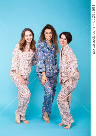 Portrait of three beautiful young girls wearing colorful pyjamas having fun during sleepover isolated over blue background. Pajama party and hen-party concept Portrait of three beautiful young girls wearing colorful pyjamas having fun during sleepover isolated over blue background. Pajama party and hen-party concept 65295691
