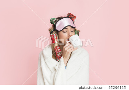Sleepy woman in curlers and sleep mask wrapped in blanket and drinks coffee from cup 65296115