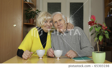 Happy old grandparents couple sit on table at home enjoy drinking tea together 65303198