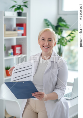 Blonde middle-aged doctor reading medcial history and looking positively Blonde middle-aged doctor reading medcial history and looking positively 65306301