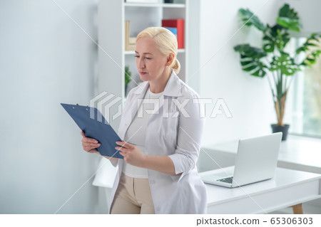 Blonde middle-aged doctor holding documents and reading Blonde middle-aged doctor holding documents and reading 65306303