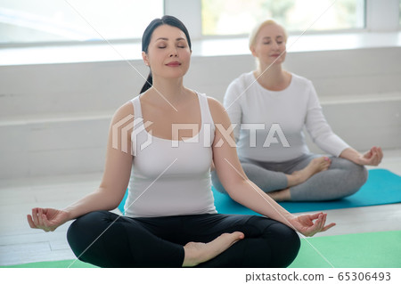 Brunette female and blonde female sitting in lotus pose, meditating 65306493
