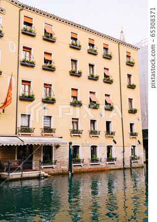 Yellow five-story building in Venice, Italy. The main entrance of the building goes into the water, at the entrance there is a sofa, on horseback there are flower pots and orange shutters from the sun 65308371