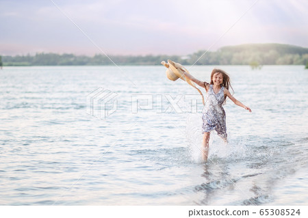 A happy young girl runs along the shore on the water and waves her hat. The girl is happy with the onset of summer 65308524