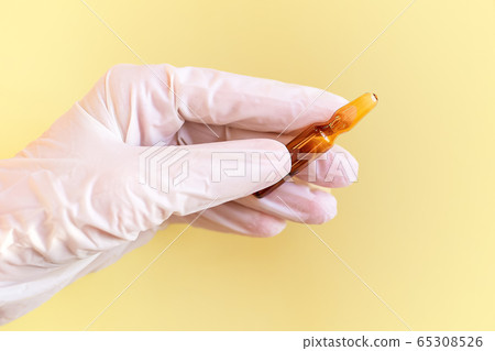 Glass ampoule in his hand. A hand in a medical glove holds the medicine on a yellow isolated background Glass ampoule in his hand. A hand in a medical glove holds the medicine on a yellow isolated background 65308526