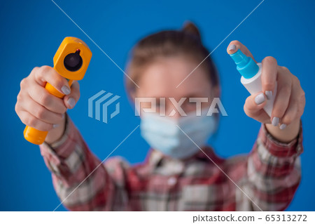 Woman holding pyrometer and spraying antiseptic toward camera against blue wall 65313272