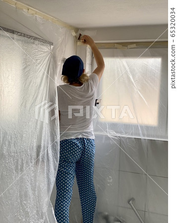 Home repairs. Woman paints in white a bathroom wall with a brush and bowl which she holds in her hands while standing on bathtub with the rest of the room covered. 65320034