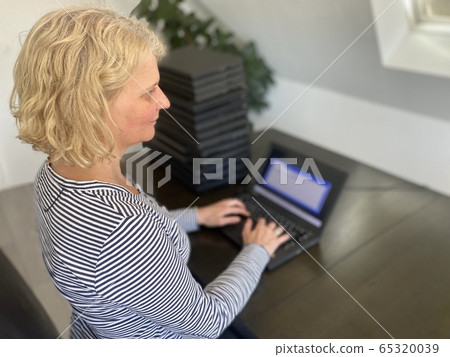 A casually dressed caucasian middle aged woman is installing a laptop computer with a stack of several more computers next to her. Laptops are used and second hand, might be for people in need due to 65320039