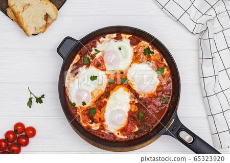 Shakshuka egg dish with tomato sauce in a black cast iron pan on the white wooden table 65323920