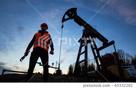 Oil man standing by pump jack under blue evening sky. Oil man standing by pump jack under blue evening sky. 65332202