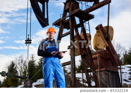 Oil engineer with a laptop standing next to an oil rig making notes in his computer Oil engineer with a laptop standing next to an oil rig making notes in his computer 65332219