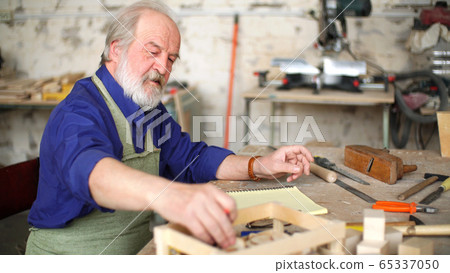 An old apprentice sits at a table in his workshop and draws a sketch of a wooden product An old apprentice sits at a table in his workshop and draws a sketch of a wooden product 65337050