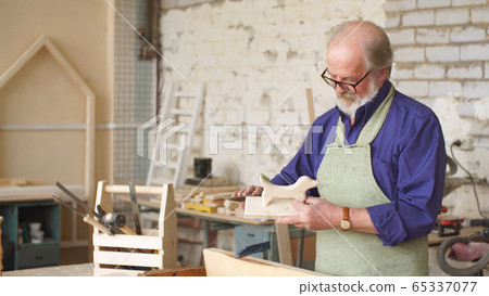 Bearded carpenter with gray hair works with wooden workpieces 65337077