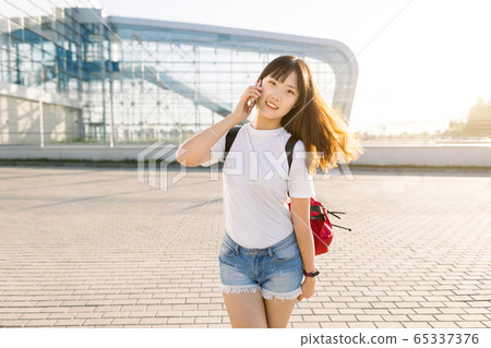 Pretty cheerful 25-aged asian brunette girl in jeans shorts and white t-shirt enjoying her conversation with friend on smartphone in front of modern airport building. Travel and technologies concept Pretty cheerful 25-aged asian brunette girl in jeans shorts and white t-shirt enjoying her conversation with friend on smartphone in front of modern airport building. Travel and technologies concept 65337376