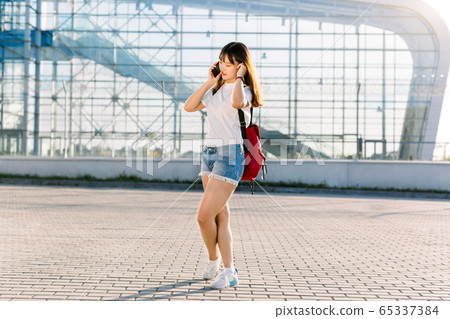 Pretty smiling asian girl in casual wear and with red backpack, talking phone with her friend while standing on sunny day in front of modern airport glass building background 65337384