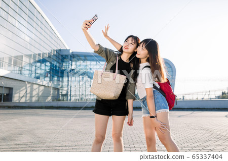 Summer lifestyle portrait of two beautiful smiling young Chinese girls in trendy clothes, which are standing near modern airport and posing on smartphone camera to make selfie photo Summer lifestyle portrait of two beautiful smiling young Chinese girls in trendy clothes, which are standing near modern airport and posing on smartphone camera to make selfie photo 65337404