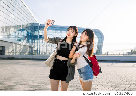 Tourism, friendship and journey concept. Two cheerful good-looking young asian girls smiling and making selfie photo on mobile phone, posing near the modern airport building before their trip 65337406