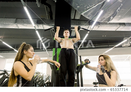 shot of a muscular Caucasian male athlete doing pull ups at the gym. Strong energetic sportsman pulling up on a bar at sport studio, showing workout for two pretty girls shot of a muscular Caucasian male athlete doing pull ups at the gym. Strong energetic sportsman pulling up on a bar at sport studio, showing workout for two pretty girls 65337714