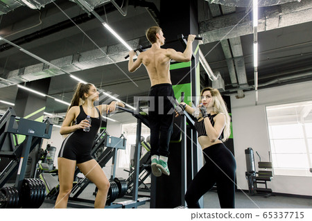 Handsome power athletic man training pumping up back muscles pull up in the gym. Strong bodybuilder trains in the gym, trains his back. Two girls with water bottles standing near bar 65337715