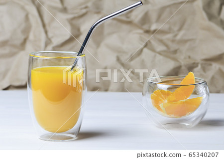 A glass of orange juice with a metal cocktail straw next to slices of orange in a transparent bowl 65340207