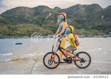 Active school kid boy and his mom in medical mask and safety helmet riding a bike with backpack on 65340423