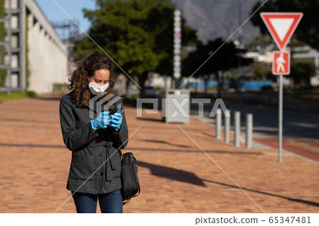 Caucasian woman wearing a protective mask and gloves out in the streets, using her phone 65347481