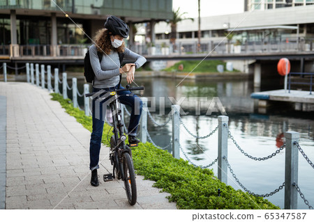 Caucasian woman wearing a protective mask and a cycling helmet in the streets, checking her smar 65347587