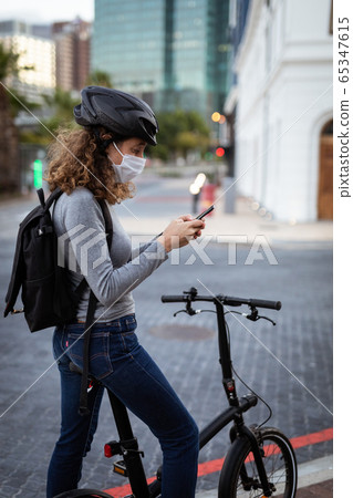 Caucasian woman wearing a protective mask and a cycling helmet, using her phone in the streets 65347615