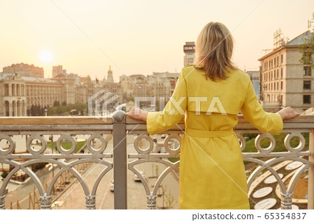 Rear view of woman looking at panorama of evening city. Adult female in yellow raincoat outdoor, copy space, golden hour 65354837