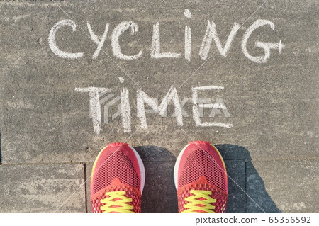Cycling time, text on gray sidewalk with woman legs in sneakers, top view 65356592