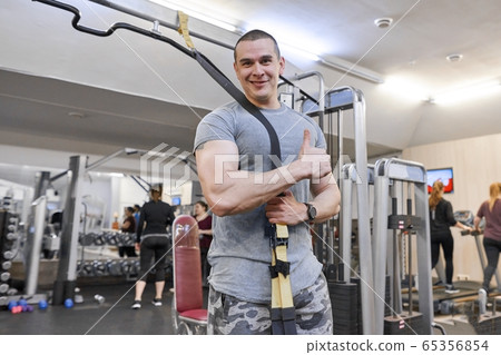Portrait of young strong smiling muscular smiling man in gym. Male with fitness straps loops Portrait of young strong smiling muscular smiling man in gym. Male with fitness straps loops 65356854
