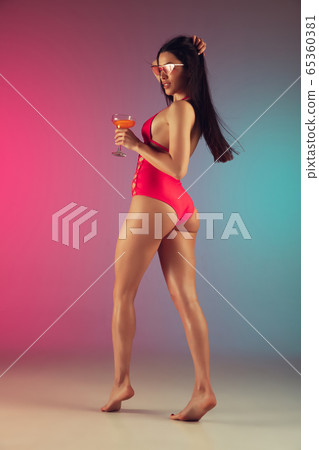 Fashion portrait of young fit and sportive woman in stylish pink luxury swimwear with cocktail on gradient background. Perfect body ready for summertime. 65360381