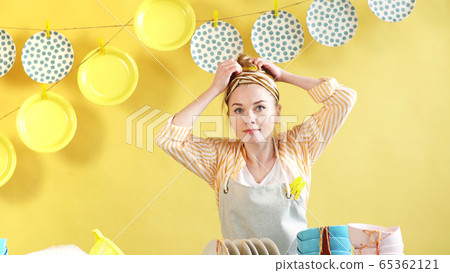 young cheerful woman put on an apron and collects her hair in a bun, before starting homework young cheerful woman put on an apron and collects her hair in a bun, before starting homework 65362121
