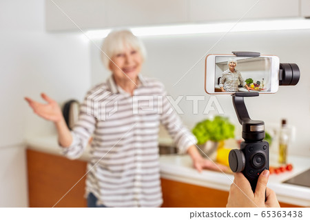 Retirement. Senior woman blogger standing at kitchen while being recorded on smartphone for culinary vlog close-up smiling friendly Retirement. Senior woman blogger standing at kitchen while being recorded on smartphone for culinary vlog close-up smiling friendly 65363438