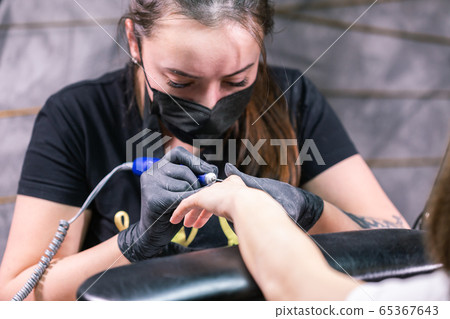 Close-up shot of hardware manicure in a beauty salon. Manicurist is applying electric nail file drill to manicure on female fingers. Close-up shot of hardware manicure in a beauty salon. Manicurist is applying electric nail file drill to manicure on female fingers. 65367643