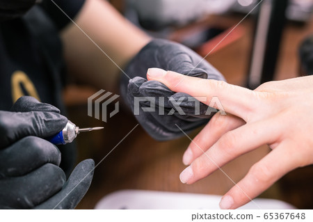 Close-up shot of hardware manicure in a beauty salon. Manicurist is applying electric nail file drill to manicure on female fingers. Close-up shot of hardware manicure in a beauty salon. Manicurist is applying electric nail file drill to manicure on female fingers. 65367648