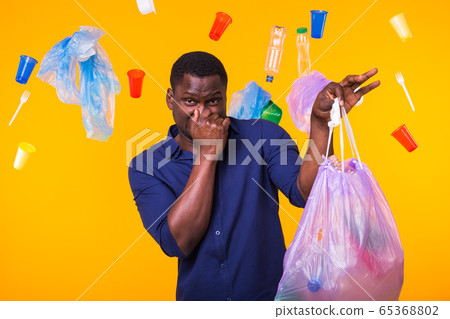 Environmental pollution, plastic recycling problem and waste disposal concept - angry african american man holding garbage bag on yellow background. He is feel smell of trash. 65368802