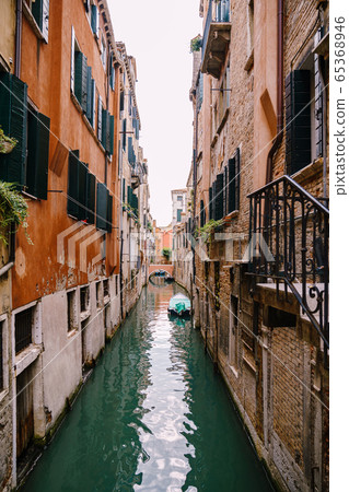 Beautiful narrow canal in Venice, Italy. Between two buildings. A small bridge in distance. Moored boat against the wall of the house. The left building is orange, the right one is made of bare brick. 65368946
