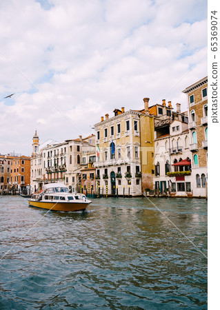 Tourists on the waterfront in Venice, against the background of the ancient facades of buildings standing in the water. Water sightseeing in Venice, Italy. Tourists on the waterfront in Venice, against the background of the ancient facades of buildings standing in the water. Water sightseeing in Venice, Italy. 65369074